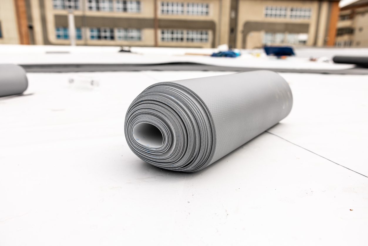 Close-up view of rolled roofing material with a backdrop of a commercial building, highlighting the comparison of TPO vs. silicone roofing for commercial roofs.